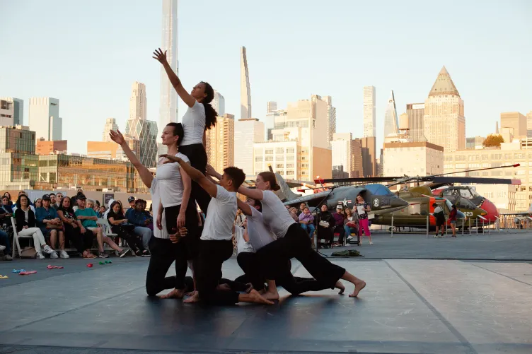 Six dancers in white shirts and black pants lift another dancer as they reach upward, with an audience, Intrepid's aircraft collection and the Manhattan skyline in the background
