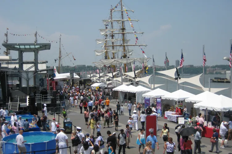 People celebrating Fleet Week on Pier 86 with tall ships docked and the Intrepid Museum nearby.