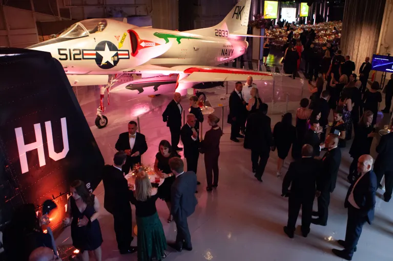 People in formal attire mingle among aircraft on the Museum's hangar deck, set with ambient lighting.