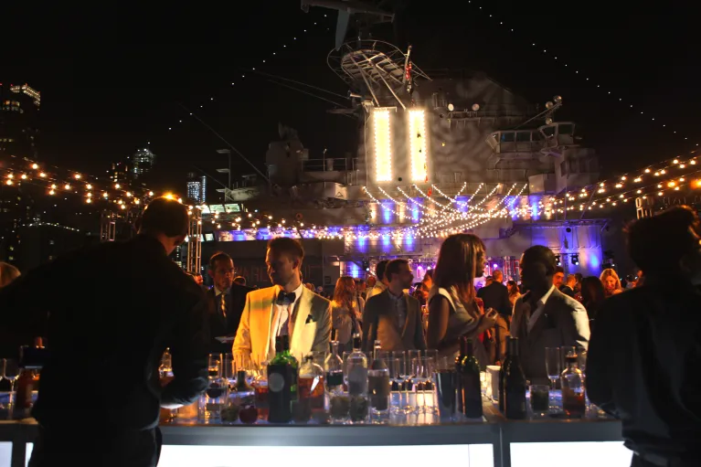 Partygoers approach a bar situated on Intrepid&#039;s flight deck at night, flanked by twinkle lights and the ship&#039;s island in the background.