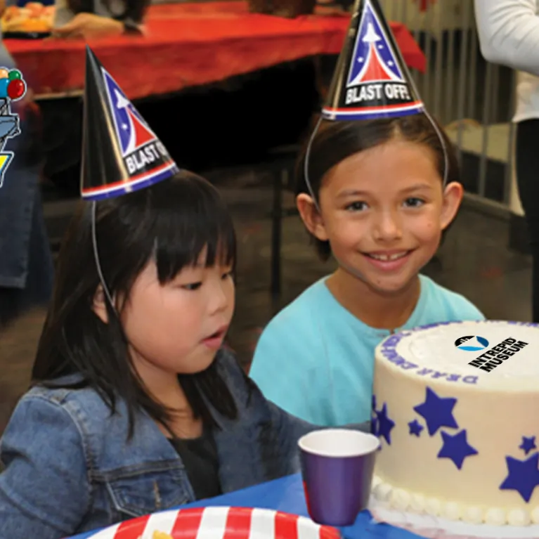 Kids wearing party hats are seated next to a large birthday cake.