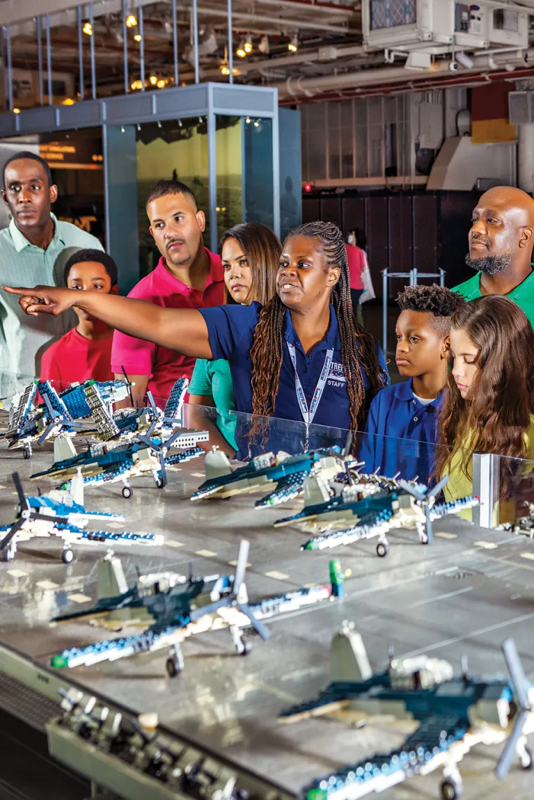 A Museum educator is with a group and pointing to the LEGO model of the Intrepid.