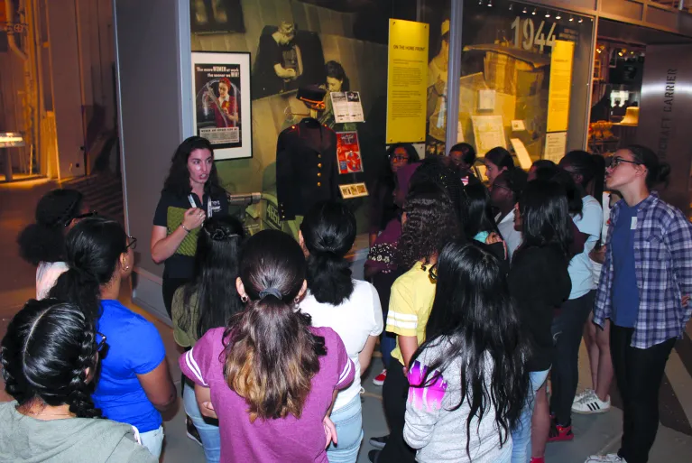 A group of visitors stand with a tour guide in front of an exhibition panel.