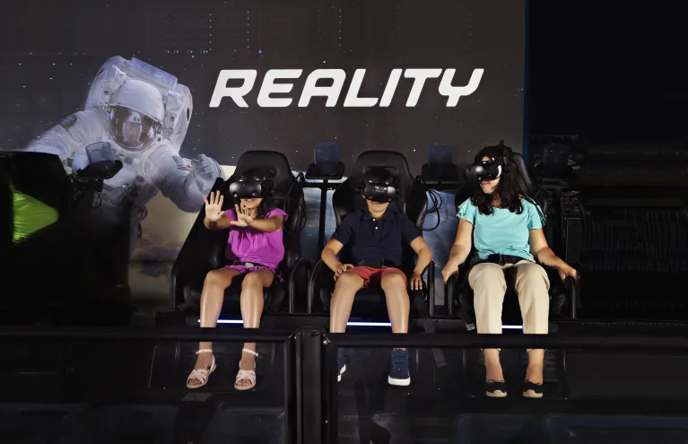 Image of a boy, woman and a girl with arm stretch out as they ride the Apollo 11 &amp; Beyond VR Experience