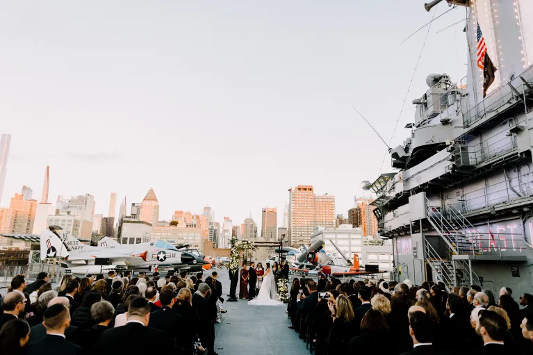 Wedding ceremony in the center of the Flight Deck, set with a chuppah and rows of black chairs with an aisle and florals