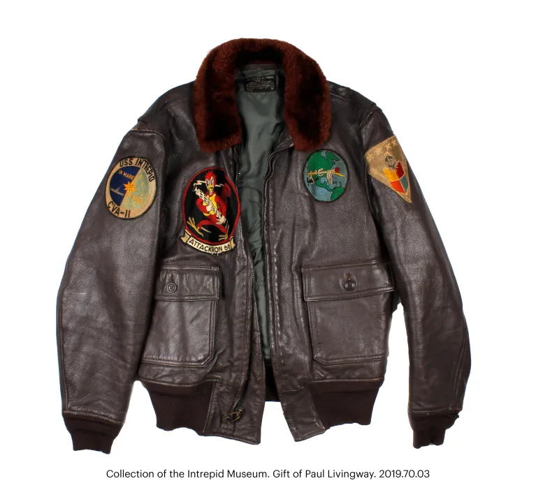 Brown leather G-1 flight or bomber jacket with a fur-trimmed collar and brown fabric waistband. It features a front zipper closure and two exterior button-flap pockets. A leather nametag is sewn on the exterior front, left side, with gold embossing depicting naval aviator wings and reading: “Oscar A. Placco // LT JG // USNR.” Three patches are sewn onto the jacket—one on each shoulder and one on the front right. A black fabric label with yellow thread on the interior collar reads: “Flight Jacket // Intermed