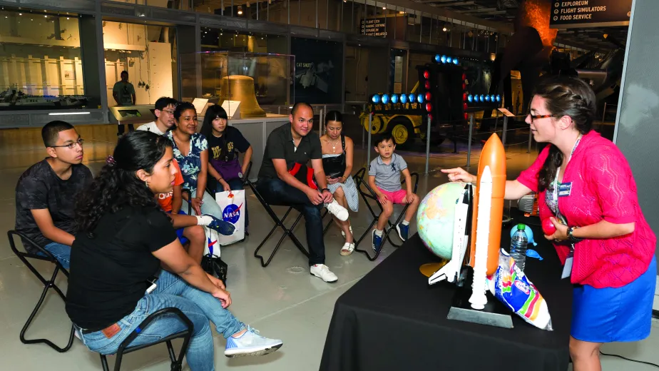 A Museum educator is standing behind a table that has a globe and a model rocket on it and is speaking to a group of seated visitors.