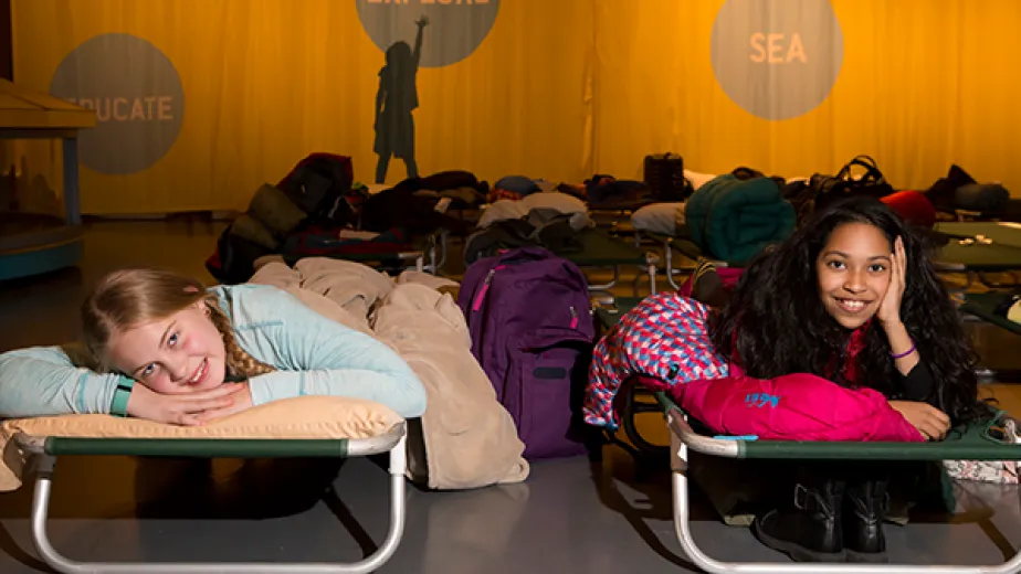 Two kids in sleeping bags are laying on top of cots on the hangar deck