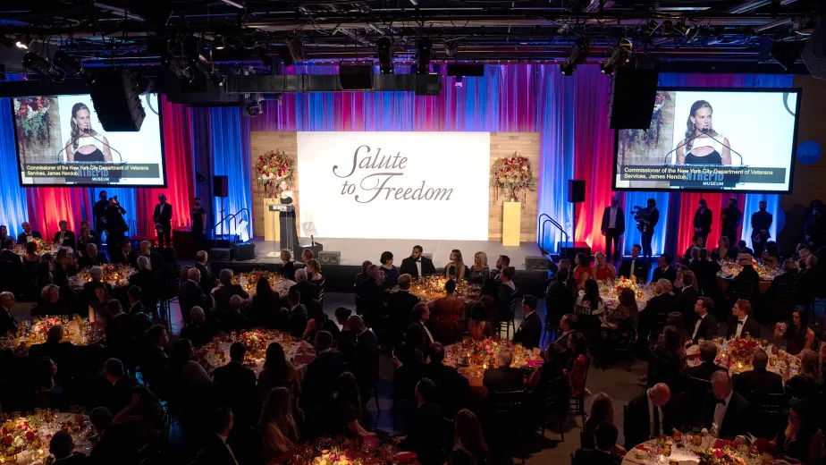 Intrepid Museum president Susan Marenoff-Zausner is speaking at a podium at the Museum's gala event with guests at tables seated nearby.