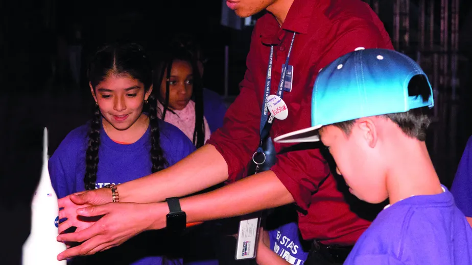 A tour guide is pointing at the nose of a space rocket in front of a group of kids.