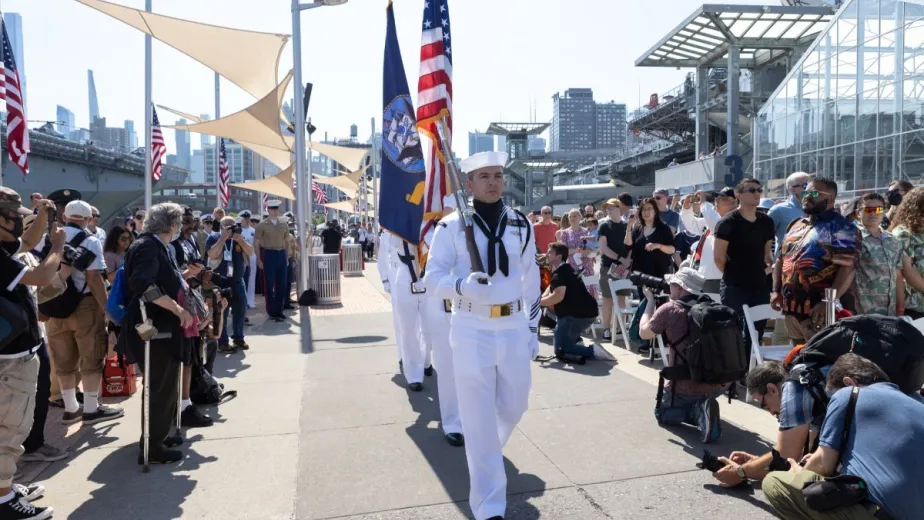 Active duty military personnel, matched and holding the American flag, are flanked by crowds on both sides.