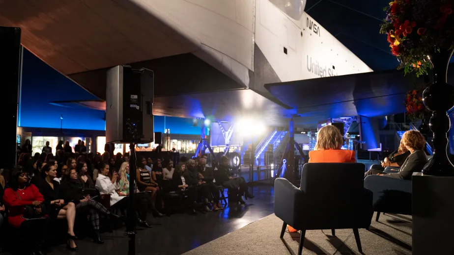 A diverse group of people seated beneath the Space Shuttle pavilion, gathered for a discussion.