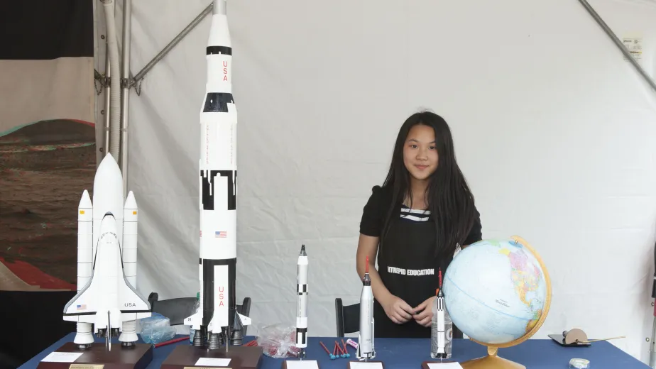 A girl is standing at a table, examining replicas of rockets and a globe.