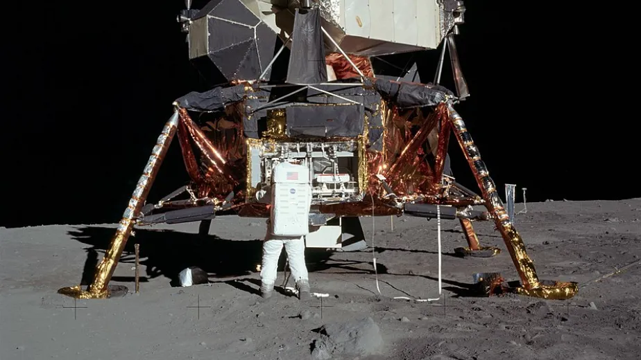 A lunar lander sits on the moon. A person in a space suit has their back to the camera, their arms on the lander.