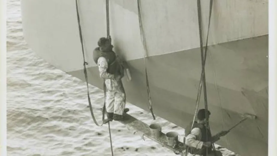 Two men are hanging by ropes from the side of a ship. They have paint cans and paint rollers.