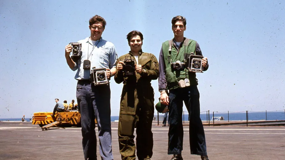 Archival photograph of three men standing on the flight deck holding cameras.