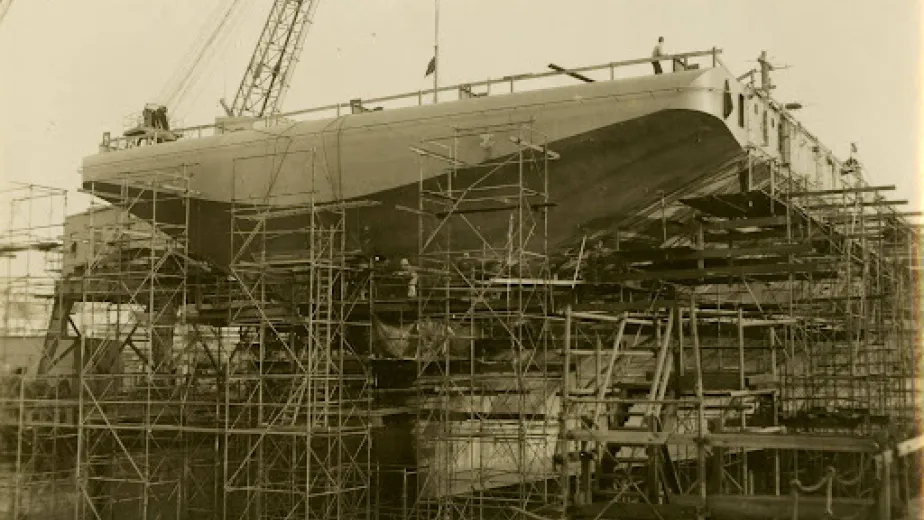Archival photograph of many scaffolding structures holding up a ship. People are standing on various parts of the scaffolding. A crane is in the background.
