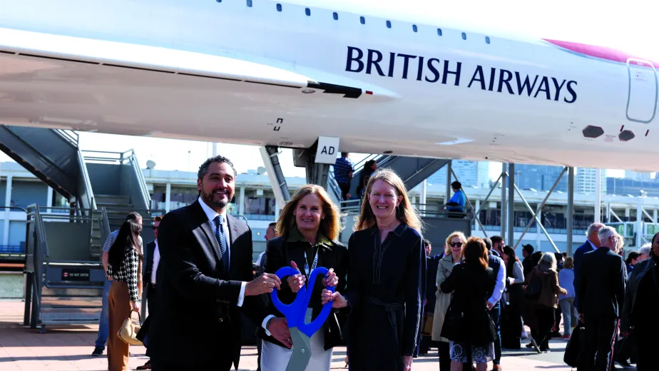 Three people standing in front of a British Airways aircraft, holding oversized scissors and a cut ribbon.