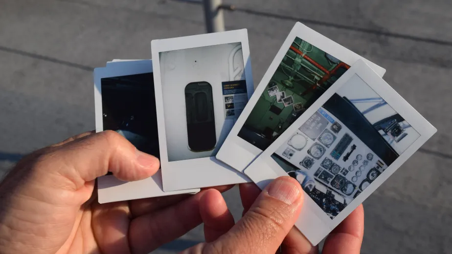 A hand holds a stack of Polaroid photos showcasing various interior views of The Intrepid, including control panels and operational spaces.