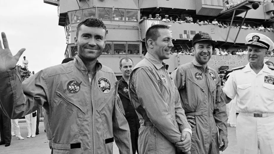 Four astronauts in flight suits, one waving, are greeted by a naval officer on the deck of an aircraft carrier, celebrated by onlookers for their achievements in space exploration.