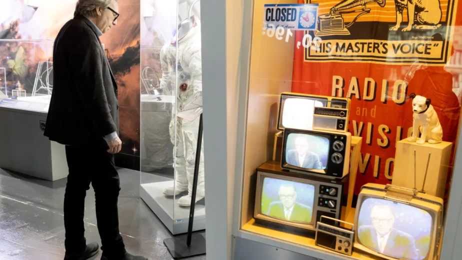 A man observing a museum display featuring a space suit and vintage televisions with a "His Master's Voice" logo and dog statue.