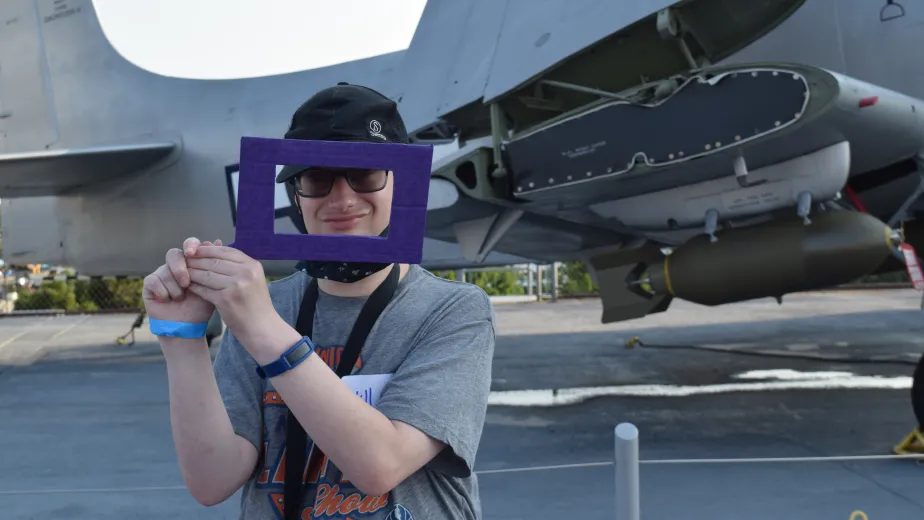 A young man wearing glasses and a black cap holds up a purple rectangular frame in front of an airplane.