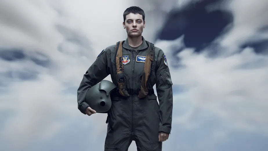 A person in a military flight suit, holding a helmet, stands confidently against a dramatic, cloudy sky.