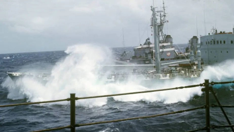 Archival image of water crashing over the bow of a ship that is pulling alongside Intrepid. The railing of Intrepid is in the foreground. The water is choppy and the sky is gray and cloudy.