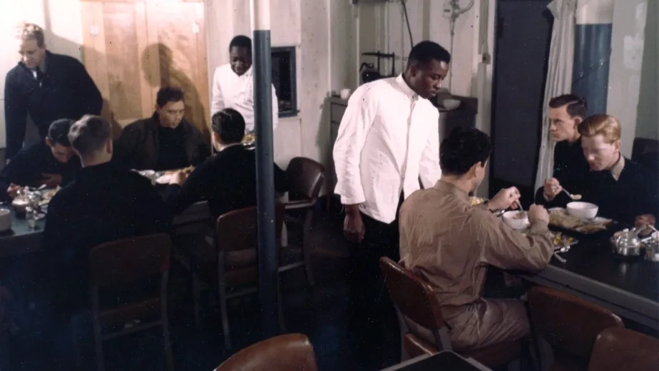 A group of men sitting at a table, enjoying a meal together, while two men stand beside the table, serving food to the group.