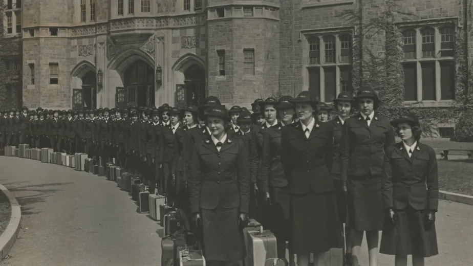  A black-and-white photo of women in uniform lined up in front of a brick building.