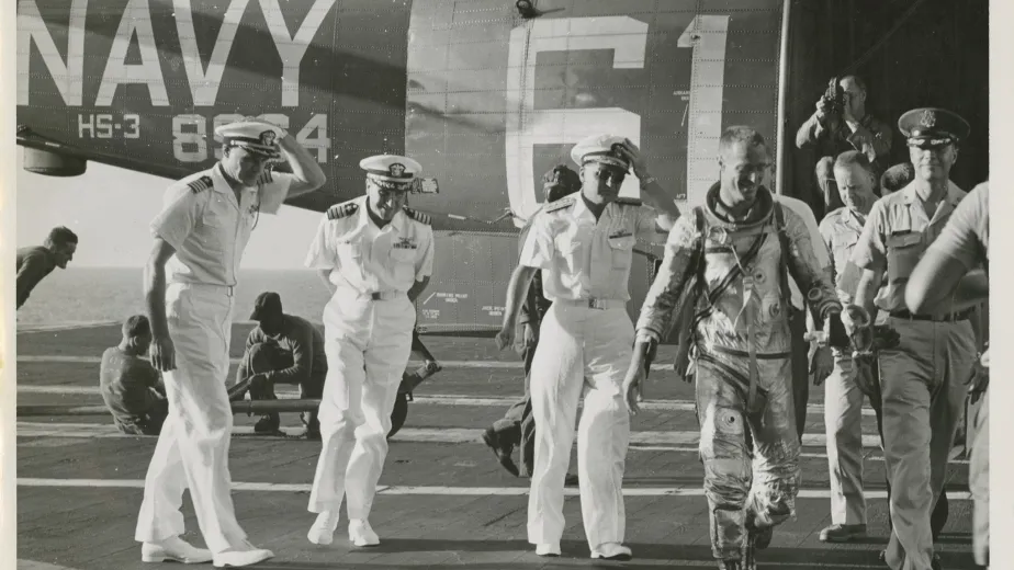 Astronaut Scott Carpenter walking out of a helicopter on a flight deck, surrounded by Naval officers.