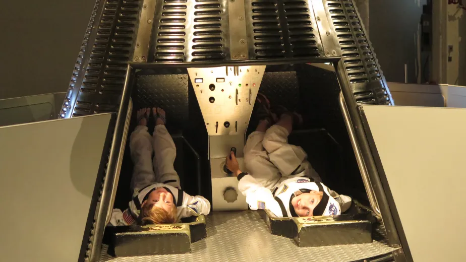 Two children in space suit costumes, sitting in a replica Gemini capsule.