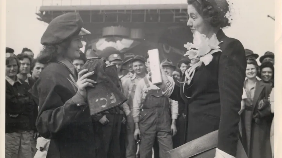 Black and white photograph showing a group of people, with two women prominently in the foreground engaged. One woman wears a uniform and cap, while the other wears a formal outfit with gloves, a large flower corsage, and a decorative hat.
