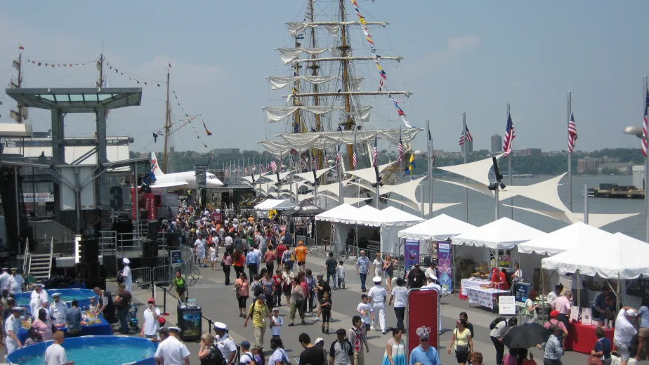 People celebrating Fleet Week on Pier 86 with tall ships docked and the Intrepid Museum nearby.