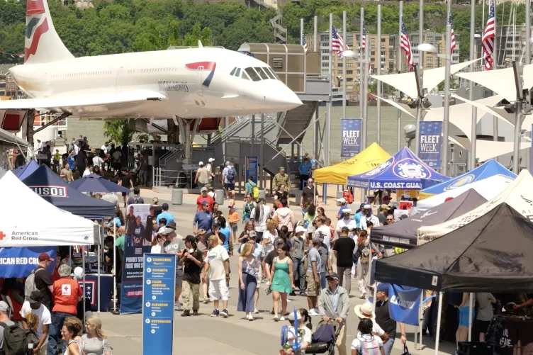 Crowds of people on Pier 86 during Fleet Week, with white event tents on the left, and active duty military personnel are sitting on military vehicles on the right.